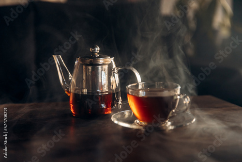 Close-up photo of a transparent teapot filled with tea and a transparent glass cup standing on a table with smoke. Hot tea for health. Smoky atmosphere for tea