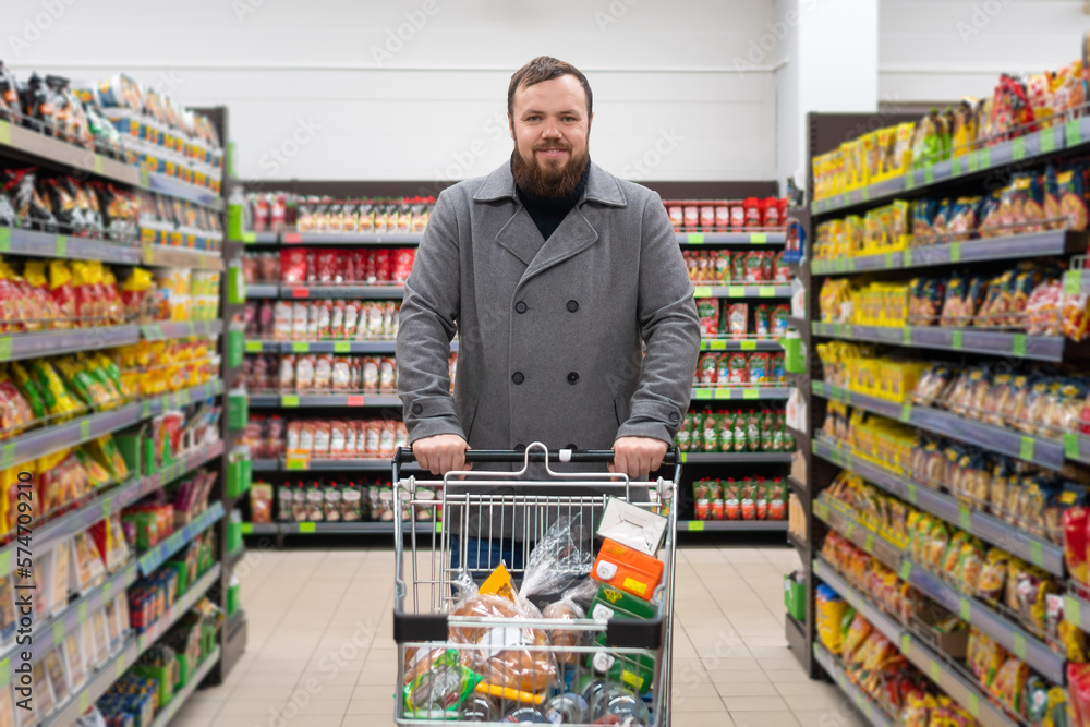 Smiling, contented young man with a beard and a gray coat is standing in a grocery supermarket with a shopping cart filled with food