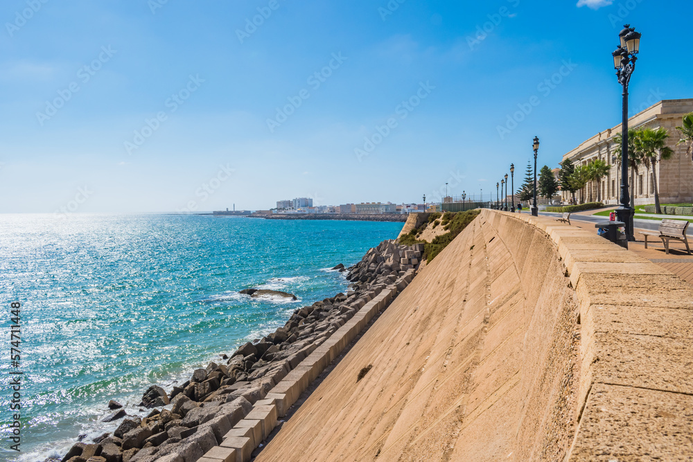 Stone wall and breakwater with blue sea and coastline of Cádiz with sunlight on horizon, SPAIN