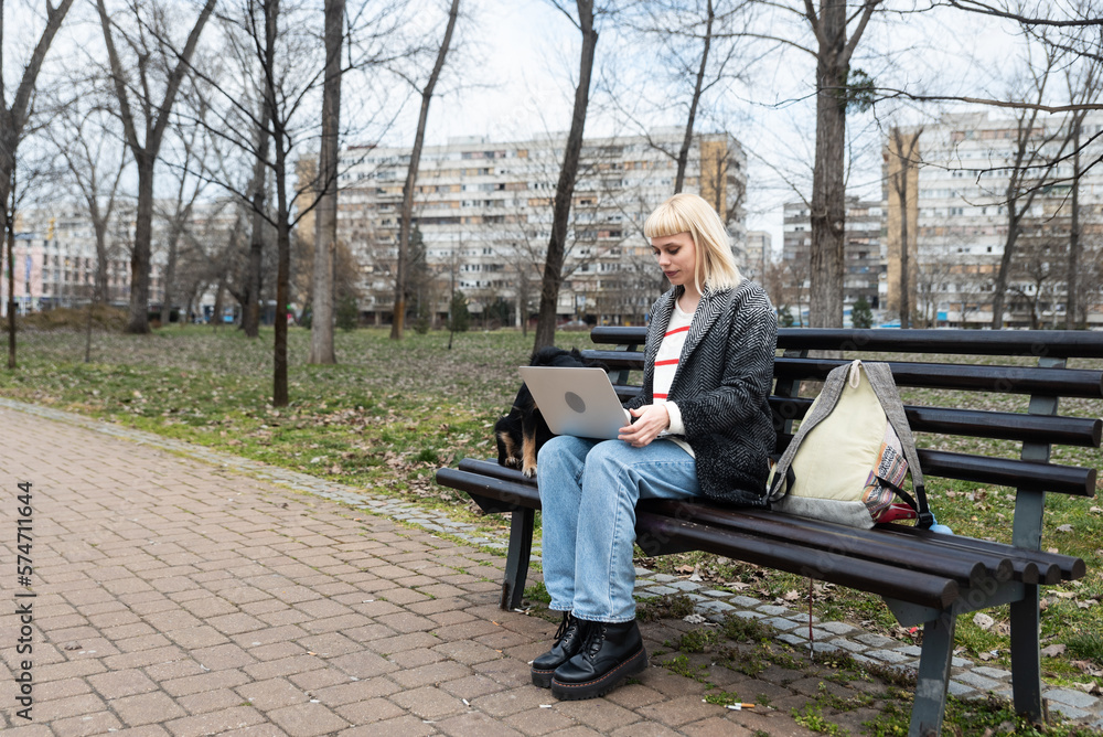 Fototapeta premium Young university girl sitting on bench with adopted dog working on laptop. She left the apartment for her student roommate boyfriend they need privacy because they share rented apartment or room