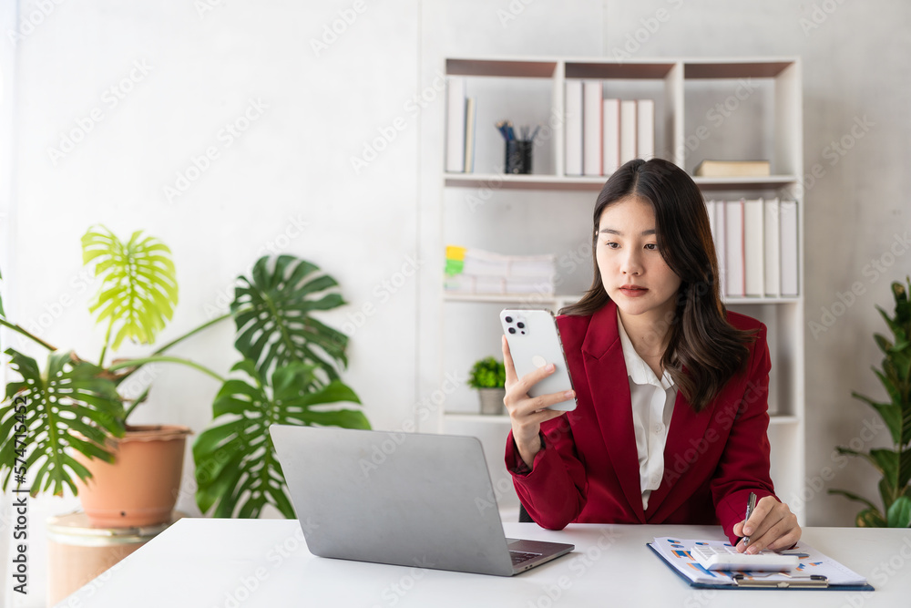 Beautiful Asian female accountant working with laptop at her desk analyzing business reports with folders, files and documents. Mobile smartphone messaging or text chatting. online work concept Beautiful Asian female accountant working with laptop at her desk analyzing business reports with folders, files and documents. Mobile smartphone messaging or text chatting. online work concept