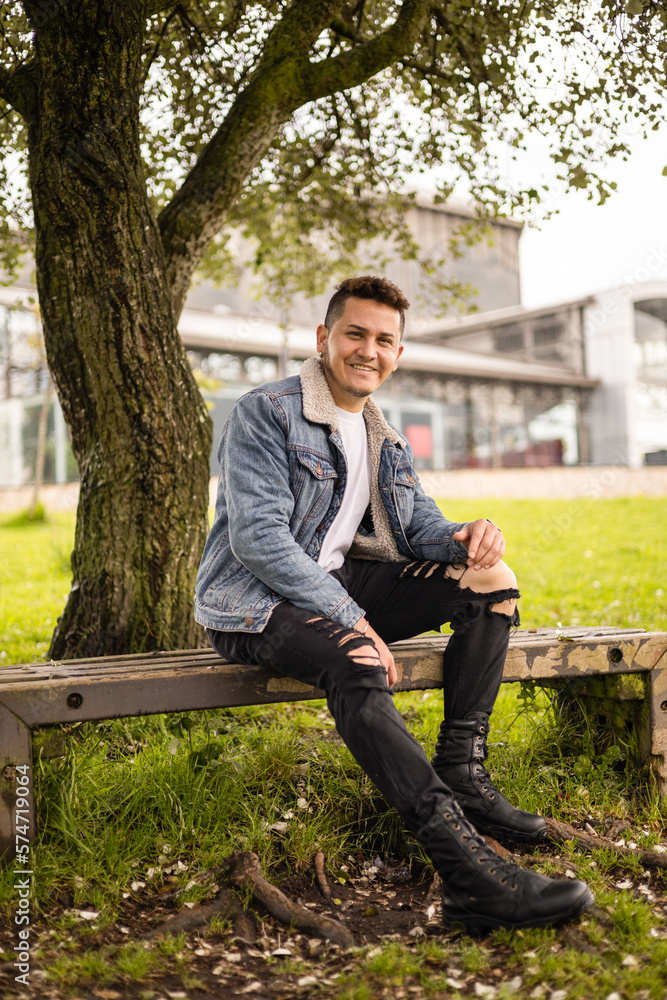 smiling young latin man resting on a park bench in the shade of a tree wears casual jean jacket, relaxed lifestyle surrounded with nature, portrait smiling young latin man resting on a park bench in the shade of a tree wears casual jean jacket, relaxed lifestyle surrounded with nature, portrait