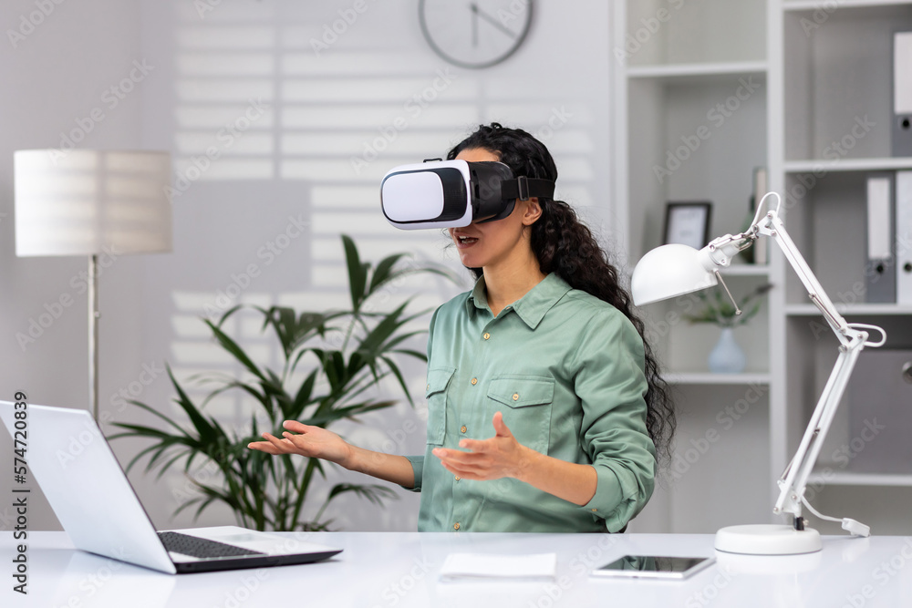 Obraz premium Successful latin american woman in office using vr glasses for video call and chatting online in virtual reality simulator, online conference with colleagues remote in metaverse.