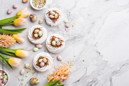 Easter treat - set of white meringues in shape of nest with multicolored chocolate eggs over marble background with tulips, hyacinths, quail eggs and sprinkles. Overhead view, flat lay, copy space