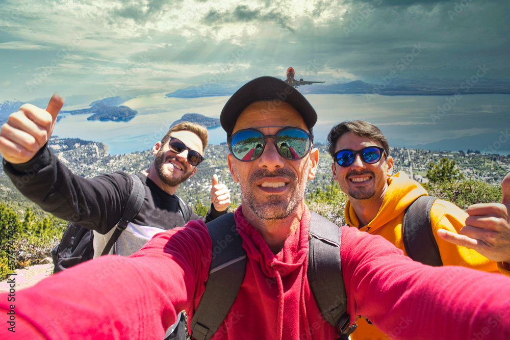 Selfie of three friends with sunglasses and backpack up Cerro Otto in Bariloche with epic view of the city, lake and mountains with cloudy skies and sunrays