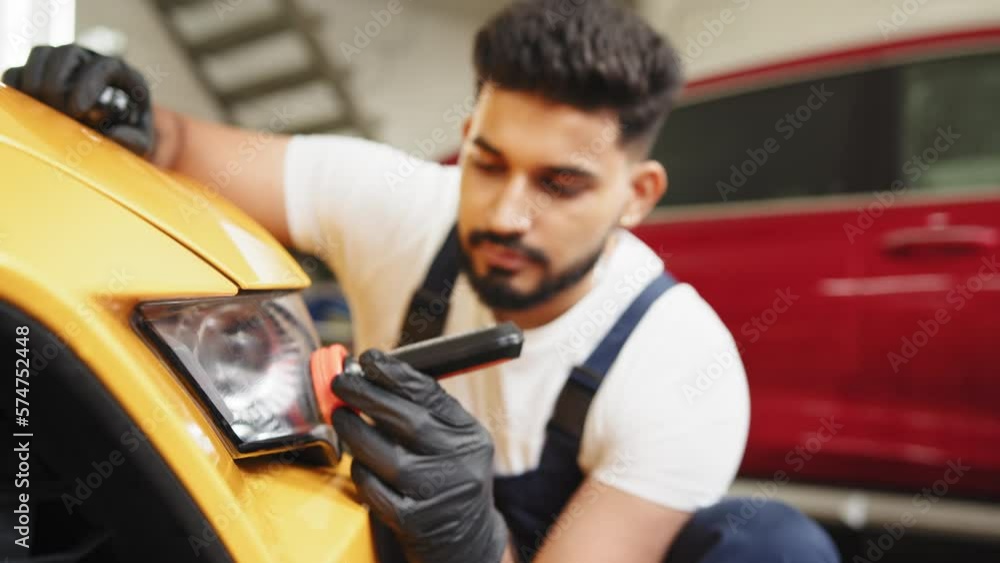 Attractive young bearded man in overalls and protective glove, applying anti-rain composition for yellow sport luxury car glass headlight in car detailing service. Attractive young bearded man in overalls and protective glove, applying anti-rain composition for yellow sport luxury car glass headlight in car detailing service.