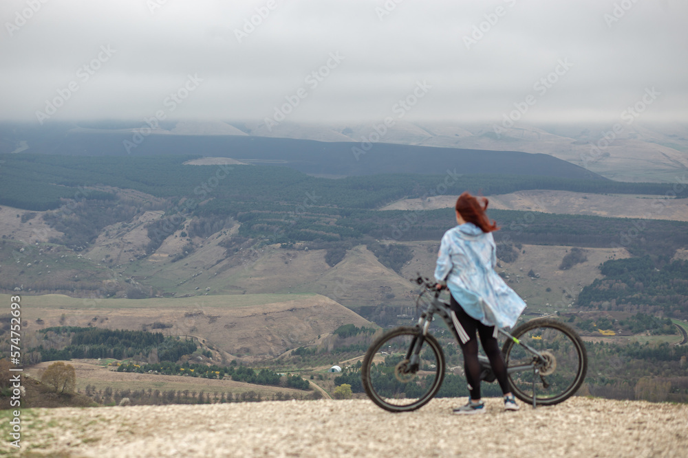 Obraz premium A woman at the top of a mountain with a bicycle looks at the admiring mountain panorama during a bicycle trip