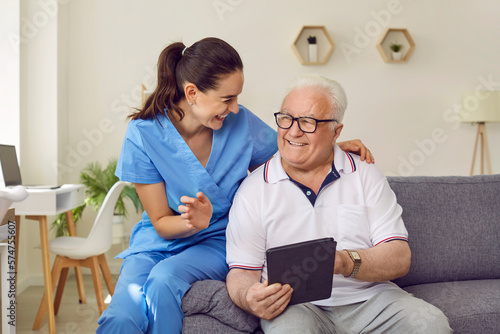 Young friendly female caregiver and senior man using digital tablet together in nursing home. Woman in medical uniform and elderly man are sitting on sofa and discussing modern technology.