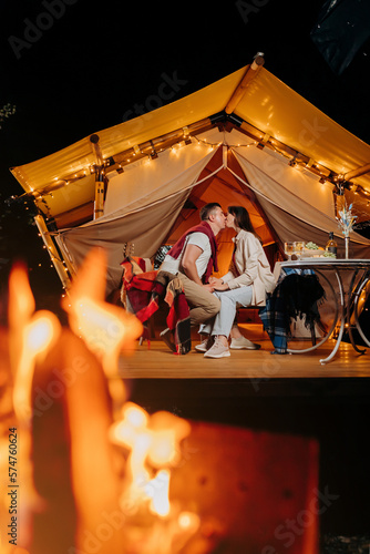 Happy lovely couple relaxing in glamping on summer evening and drinking wine near cozy bonfire. Luxury camping tent for outdoor recreation and recreation. Lifestyle concept
