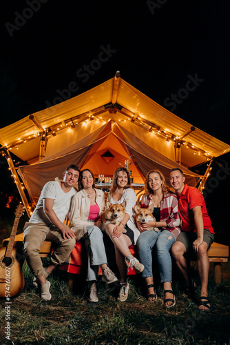 Happy group of friends with Welsh Corgi Pembroke dogs relaxing in glamping on summer evening near cozy bonfire. Luxury camping tent for outdoor recreation and recreation. Lifestyle concept © bondvit
