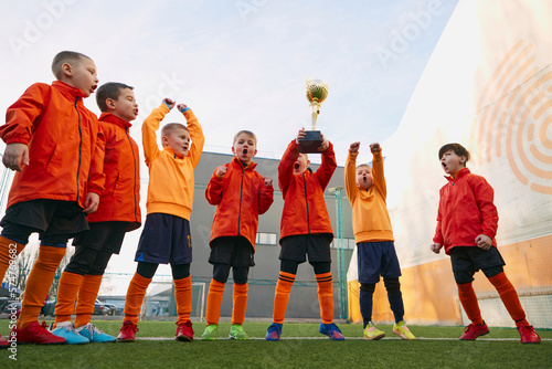 Happiness. Group of little boys, children in uniform, football players raising award, trophy. Kids training on outdoor playground. Concept of sport, childhood, active lifestyle, hobby, sport club
