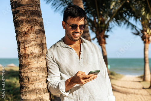 Photography Smiling man using smartphone on tropical beach under shade of palm trees