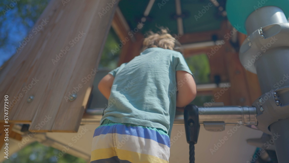 Fototapeta premium Little boy climbing playground structure outside during bright sunny day. Effortful kid going up in rope ladder outdoors. Active small kid