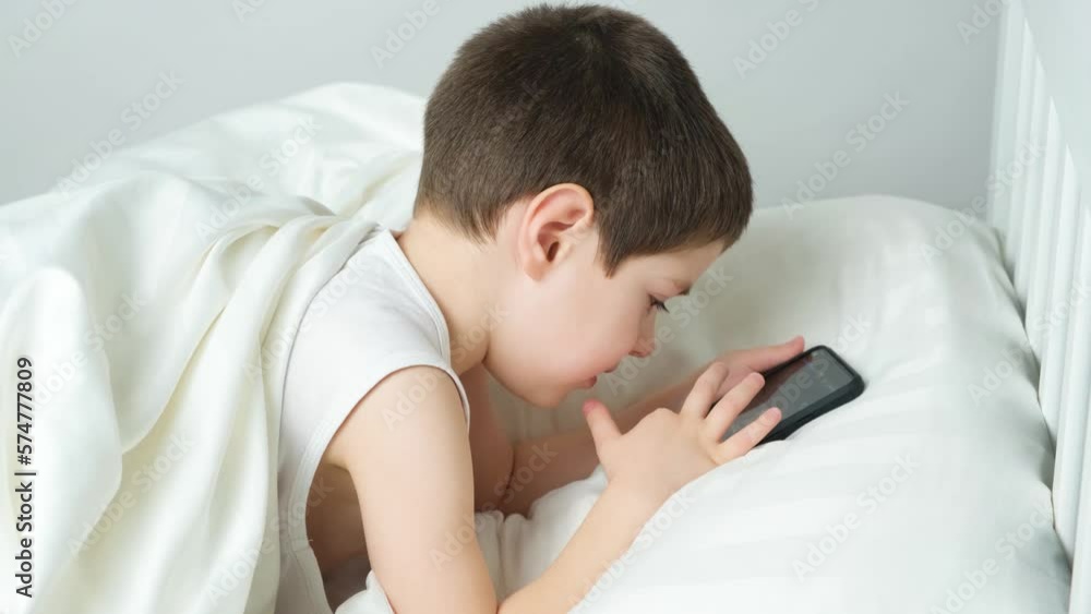 A 5 year old boy plays games on his phone while lying in bed. Gadgets in bed before bedtime in a child A 5 year old boy plays games on his phone while lying in bed. Gadgets in bed before bedtime in a child