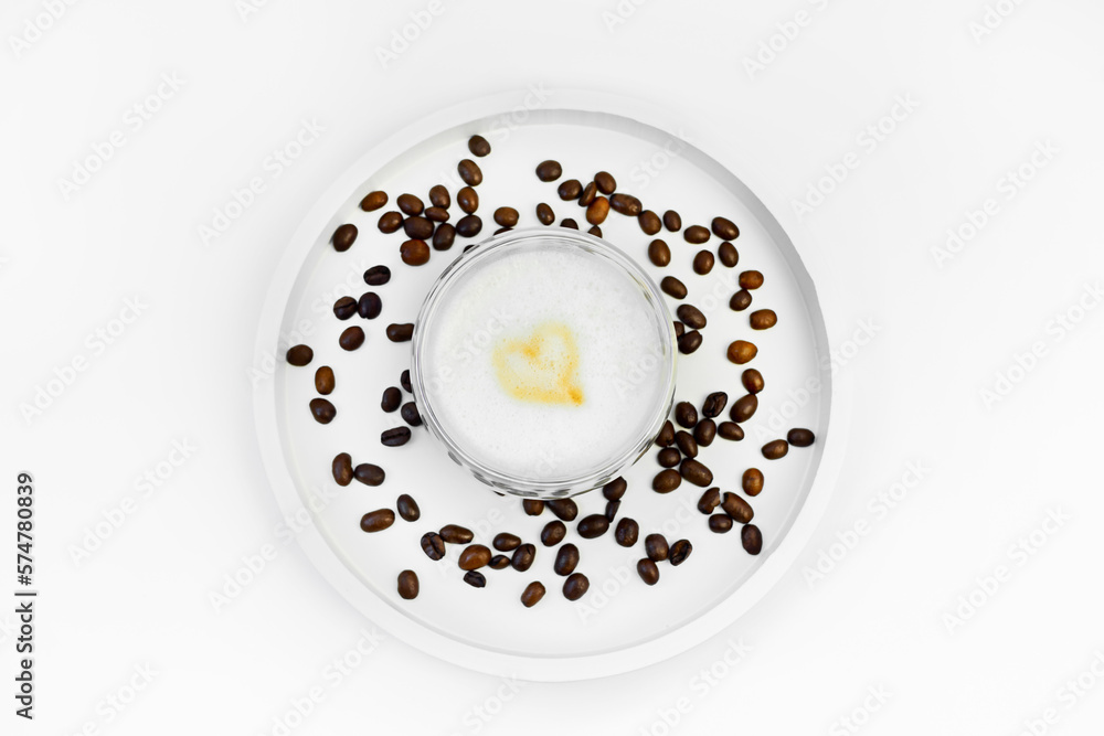 Cappuccino with a heart on the foam in a glass cup on a white stand with scattered coffee beans. Coffee view from above. Cappuccino with a heart on the foam in a glass cup on a white stand with scattered coffee beans. Coffee view from above.