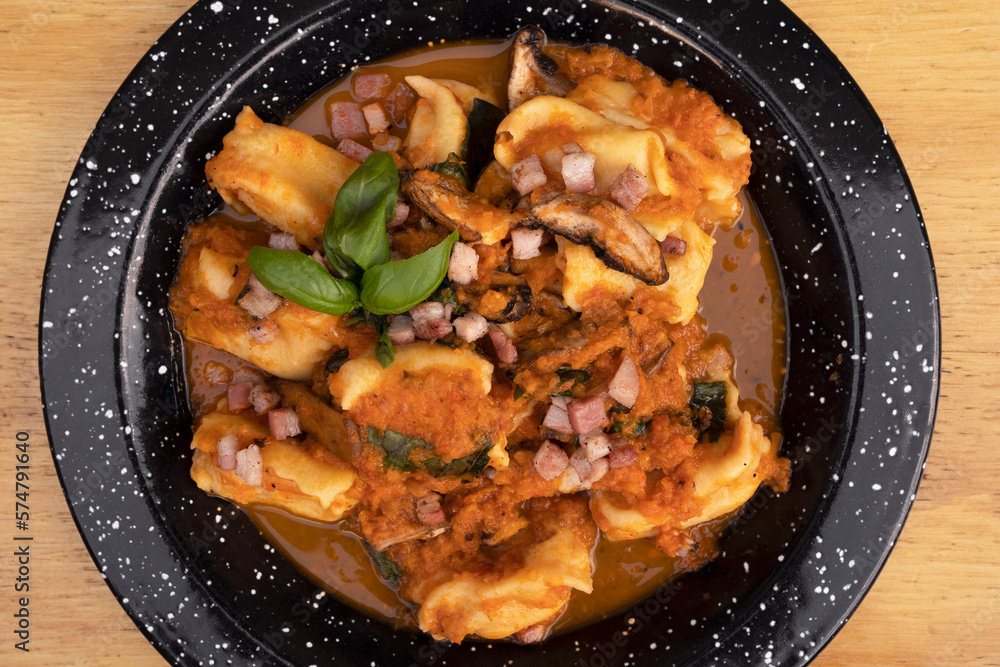 Pasta. Top view of traditional stuffed angelottis with a tomato sauce with mushrooms, bacon and basil in a black dish on the wooden table. Pasta. Top view of traditional stuffed angelottis with a tomato sauce with mushrooms, bacon and basil in a black dish on the wooden table.