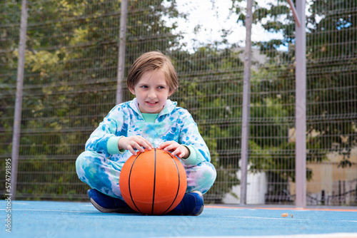 a serious little boy sits on the floor of a sports field, holds a basketball ball, watches the game