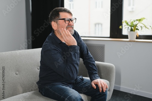 Dental Care, Tooth Sensitivity Concept. Portrait of unhappy mature man suffering from terrible toothache, touching cheek feeling acute pain, aged male sitting on the couch at home, free copy space