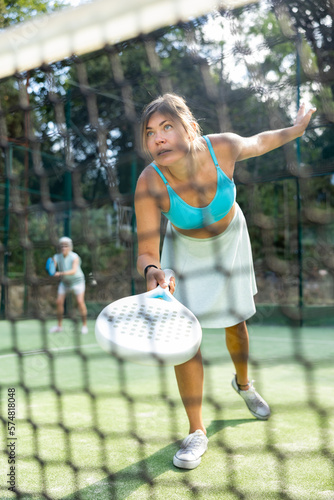 View through net of concentrated sporty young woman playing paddle tennis on outdoor court on sunny summer day, preparing to hit ball..