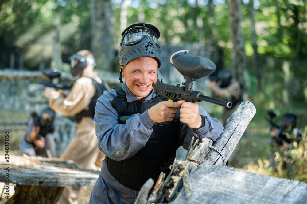 Fototapeta premium Excited caucasian man paintball player standing at cover, aiming with paintball gun and shooting.