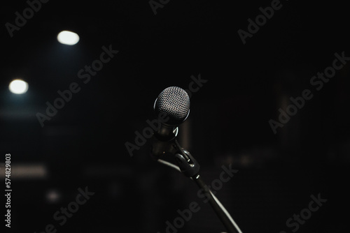 Front view of a single microphone in the middle of a dark background. Singer POV in front of a microphone on stand. Selective focus on a silver mic on stage with a lot of copy space.