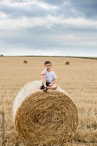 A cute little boy in a white t-shirt and khaki shorts is sitining on round bales of hay. Photo session in the field