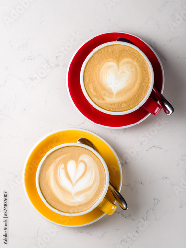 Two Cappuccino coffee with heart drawing at yellow and red cup on white marble table top view vertical