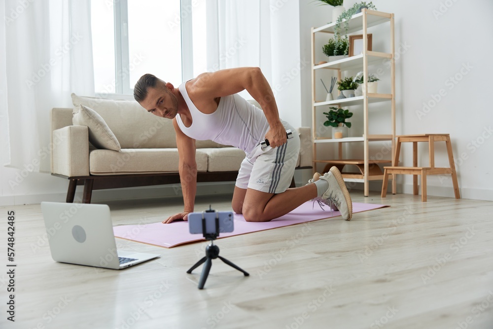 Man sports, watching a workout tape on his phone and repeating exercises sports blogger with dumbbells, pumped up man fitness trainer works out at home, the concept of health and body beauty Man sports, watching a workout tape on his phone and repeating exercises sports blogger with dumbbells, pumped up man fitness trainer works out at home, the concept of health and body beauty