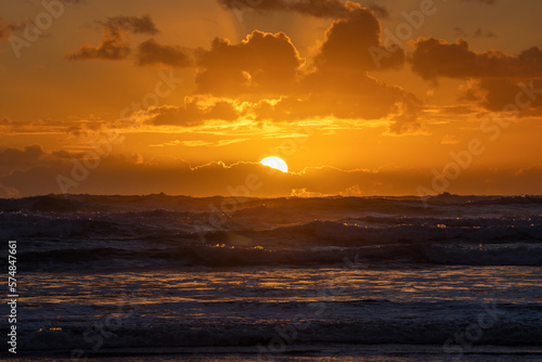 A dramatic orange sunset peeking out from behind a layer of clouds, casting a warm glow over the restless waves of the ocean. (ID: 574847661)