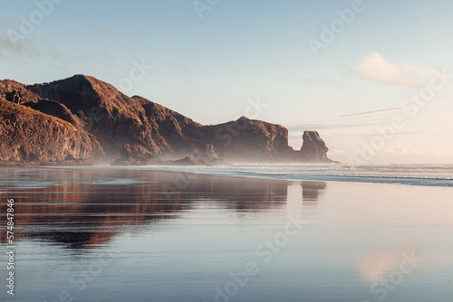 Piha beach captured at sunset, with the warm colors of the sky reflecting on the tranquil ocean waves. (ID: 574847846)