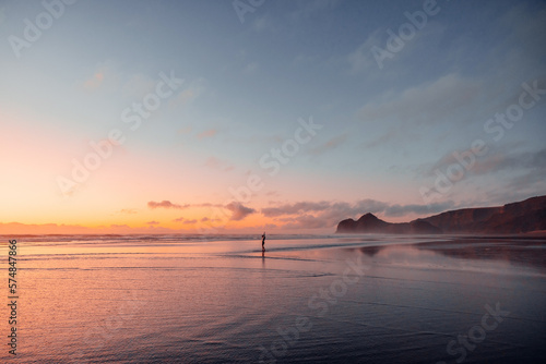 A young woman dances at Bethells beach at sunset, surrounded by vibrant pink, violet, and orange tones. (ID: 574847866)