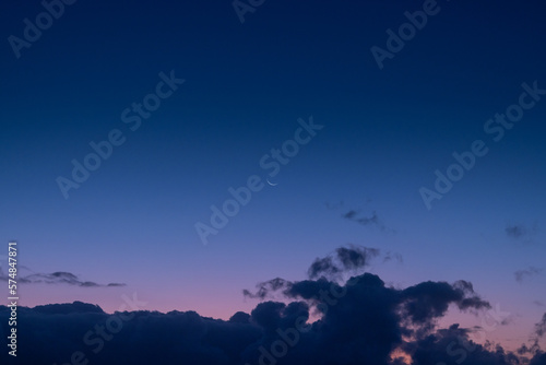 The tranquil beauty of a small crescent moon suspended in a sea of blue and purple hues, peeking out from above the rolling clouds. (ID: 574847871)