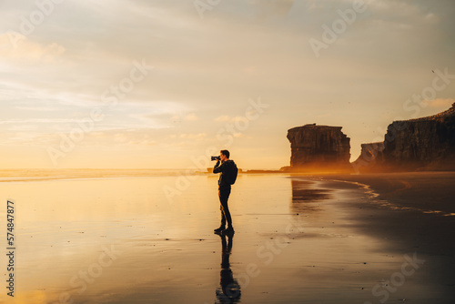 A lone photographer silhouetted against a blazing golden sunset, intently capturing the breathtaking scenery through his lens, at a stunning and secluded beach location. (ID: 574847877)