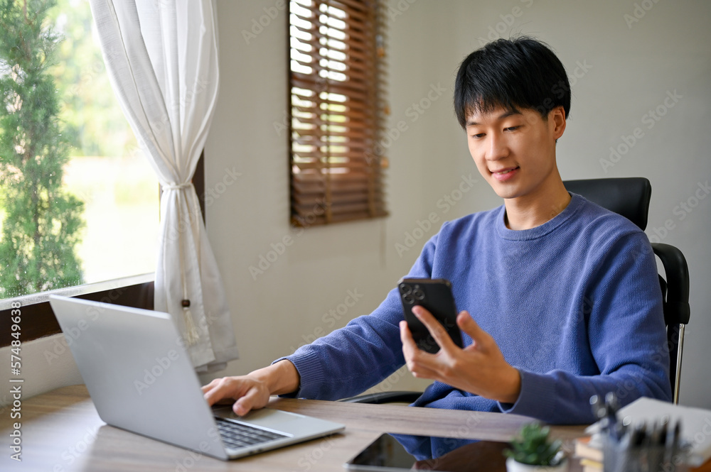 Fototapeta premium Handsome young Asian male office worker using his smartphone at his office desk.