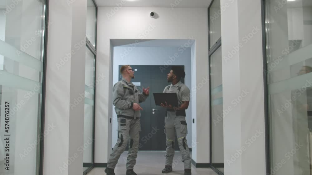 Two diverse men in uniform walk corridor and discuss CCTV cameras installation in business office. Installers set up security cameras using computer program on laptop. Concept of surveillance system. Two diverse men in uniform walk corridor and discuss CCTV cameras installation in business office. Installers set up security cameras using computer program on laptop. Concept of surveillance system.