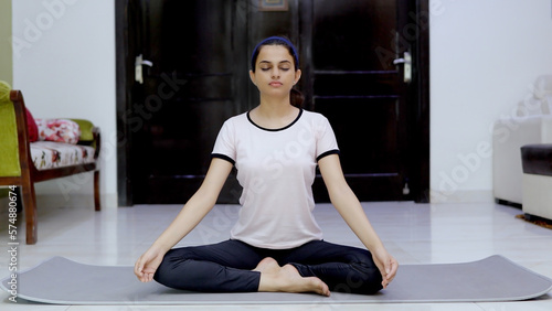 Beautiful young female doing rhythmic breathing exercises on a grey yoga mat. Attractive Indian girl meditating in Ardha Padmasan (half-lotus pose) with closed eyes - healthy lifestyle concept