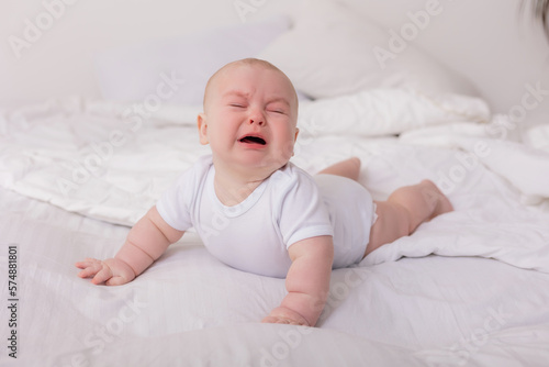 baby in a white bodysuit lies on his stomach in bed and cries