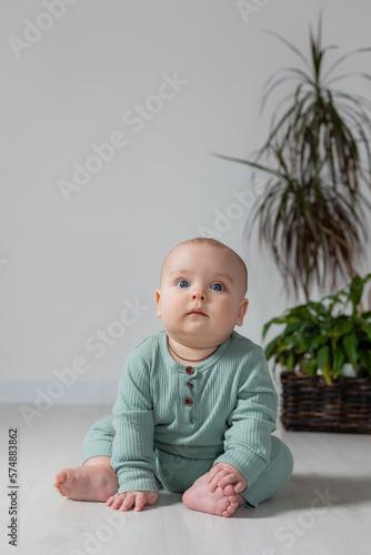 cute chubby-cheeked baby in a green jumpsuit is sitting on the floor next to a houseplant