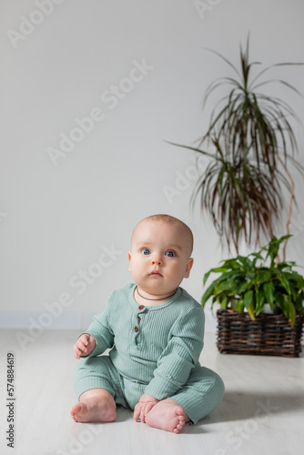 cute chubby-cheeked baby in a green jumpsuit is sitting on the floor next to a houseplant