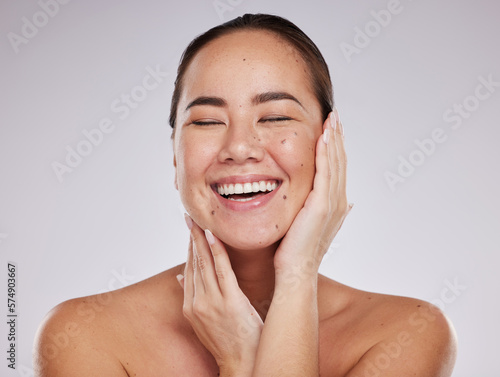 Skincare, hands on face and woman laughing in studio for cosmetics, dermatology and beauty glow. Happy aesthetic asian model person with makeup, skin mole and facial self care on grey background