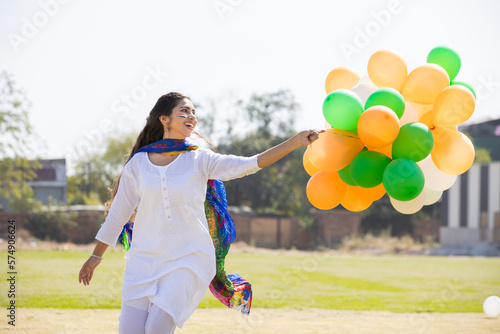 Beautiful happy young indian girl wearing traditional white kurta dress running with tricolor balloons at park celebrating independence day or republic day concept.