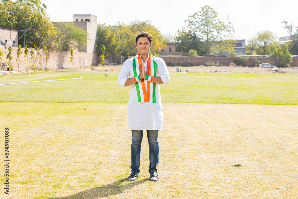 Portrait of young indian man wearing traditional white kurta and tricolor duppata do namaste while standing at park. Election and politics, celebrating Independence day or Republic day. Full length