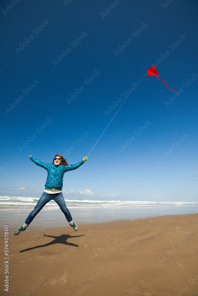 A young woman jumps into the air while flying a red kite in the sky at a beach along the Oregon Coast.