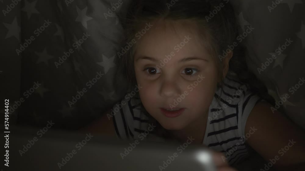 Child in dark room under covers looks at screen of tablet.girl of Internet plays online games on site.Internet security technology of future.child is playing game on Internet. Tablet device for child Child in dark room under covers looks at screen of tablet.girl of Internet plays online games on site.Internet security technology of future.child is playing game on Internet. Tablet device for child