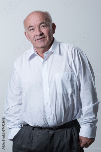 Portrait of an older man wearing a white shirt on a gray background. A healthy old man looks at a camera isolated over a gray wall. An older man smiles at the camera, a happy old man. 