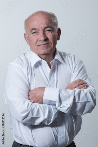 Confident senior man in white shirt crossing hands on chest and looking at camera while standing against gray background. Self confident senior isolated white studio shoot.