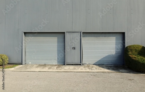 Two metallic roller shutter garage doors on a modern gray facade. Cement sidewalk and asphalt road in front. Background for copy space.