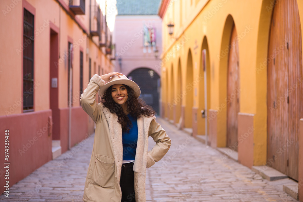 Young and beautiful Hispanic brunette woman with curly hair wearing a hat and coat for the cold walking in the city of seville while making different expressions and having fun.