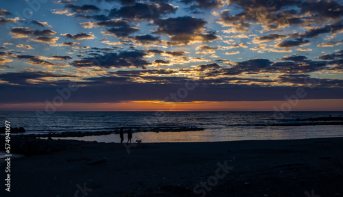 A young couple of lovers with the dog, take a romantic walk on the beach of Ostia, in Rome, in the sunset. The sun covered by clouds and the reflection of warm light on the blue sea water.