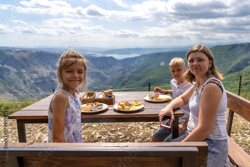 Happy mother with teenage son and daughter are sitting at wooden table on cliff and having breakfast with view of picturesque Sulak Canyon in Dagestan Perfect breakfast with pancakes and mountain view © sommersby